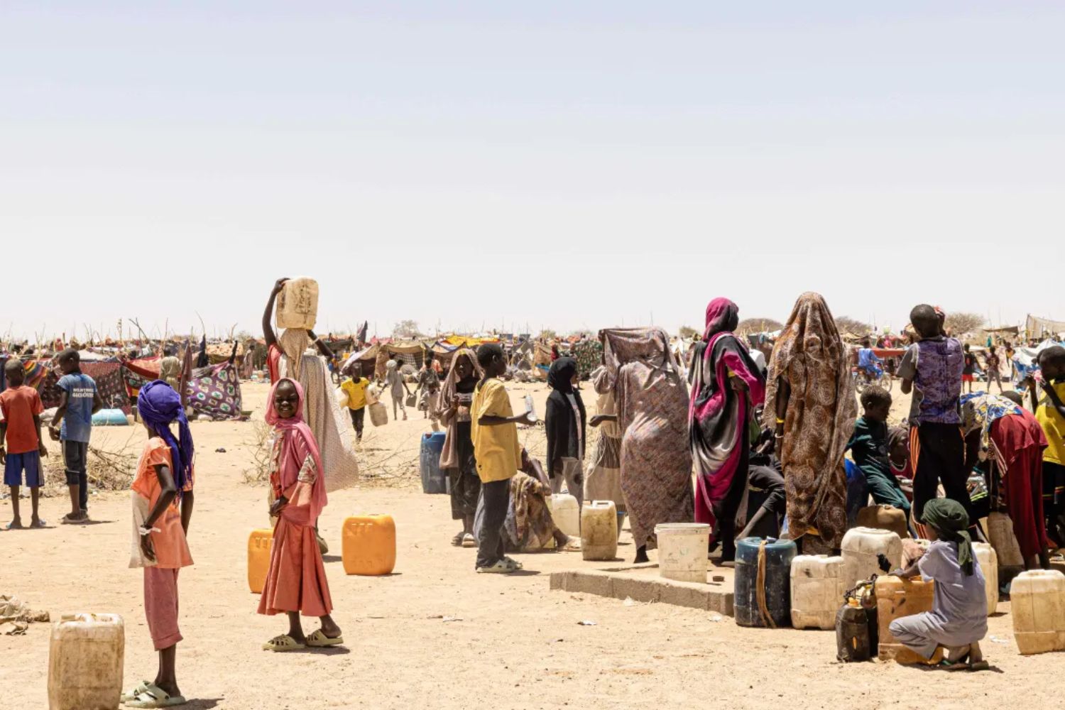 People gather in a sandy area, carrying water containers and wearing colorful clothing, with a backdrop of makeshift shelters and a clear sky.