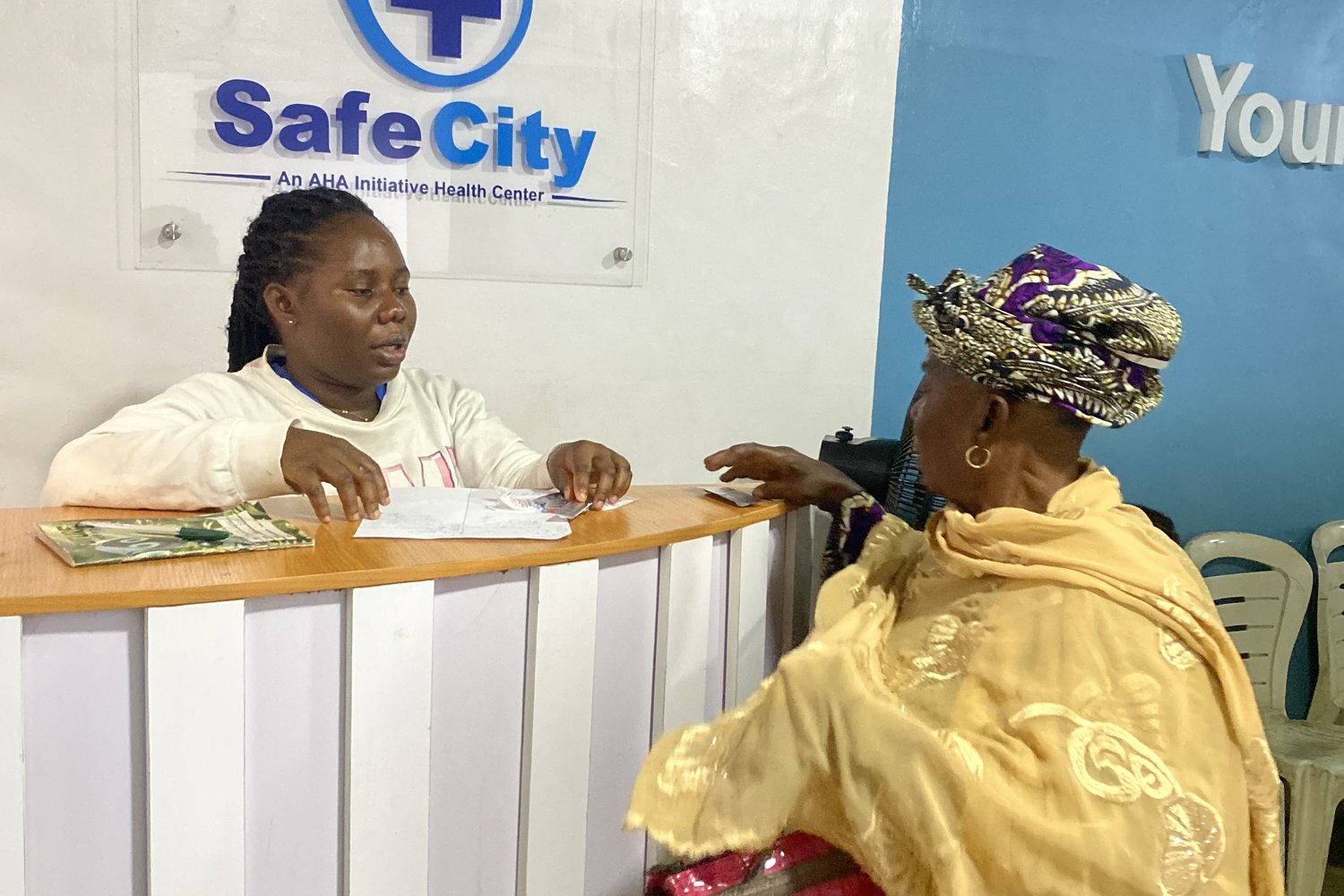 Woman in gold embroidered dress and patterned headwrap speaking with a receptionist at the Safe City Health Centre front desk.