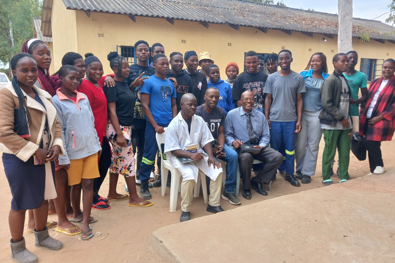 A group of people, including two seated men, pose for a photo outside a building with a corrugated metal roof.