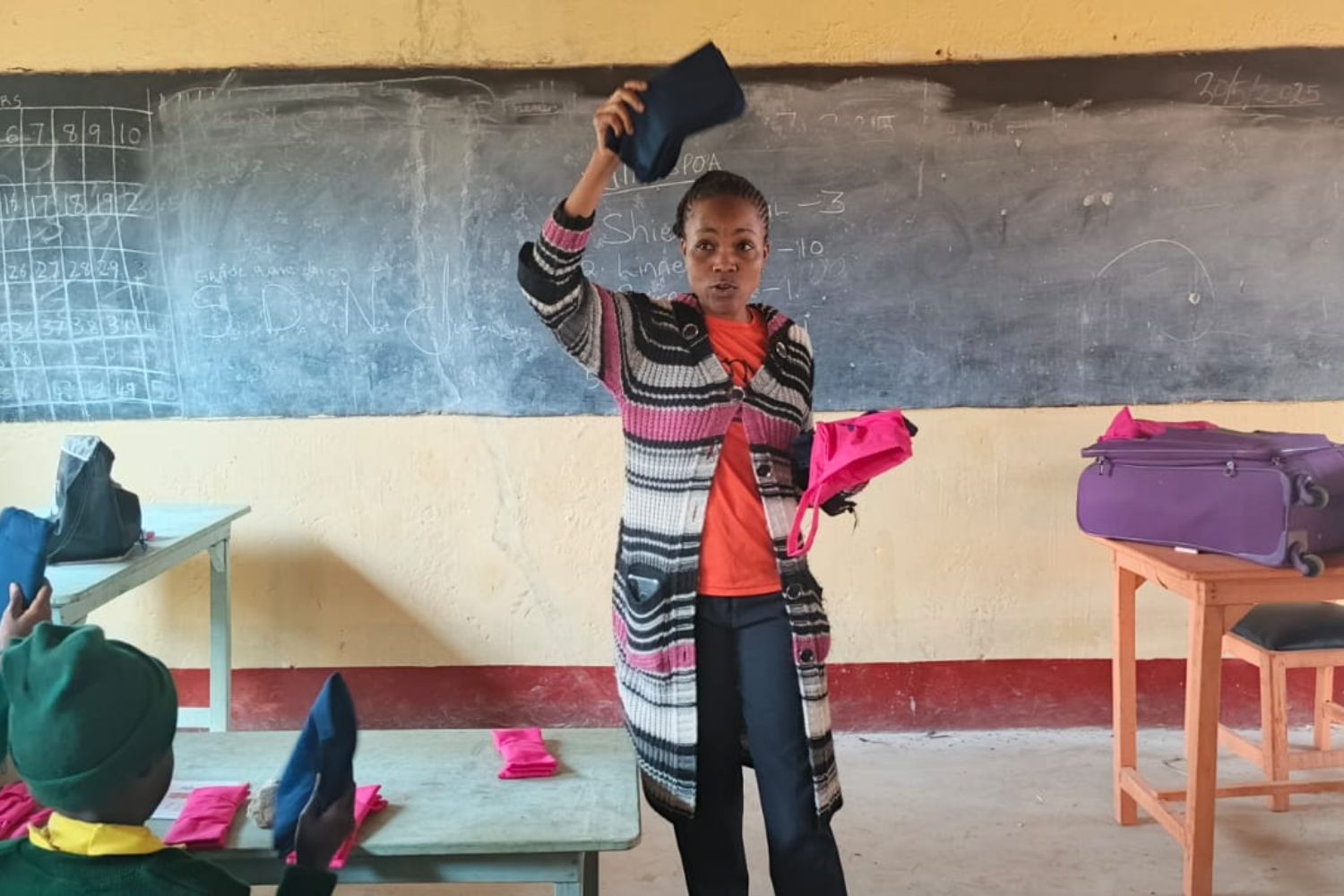 A teacher in a classroom holds up a blue item, possibly a bag, while distributing pink items to seated students. A chalkboard is in the background.