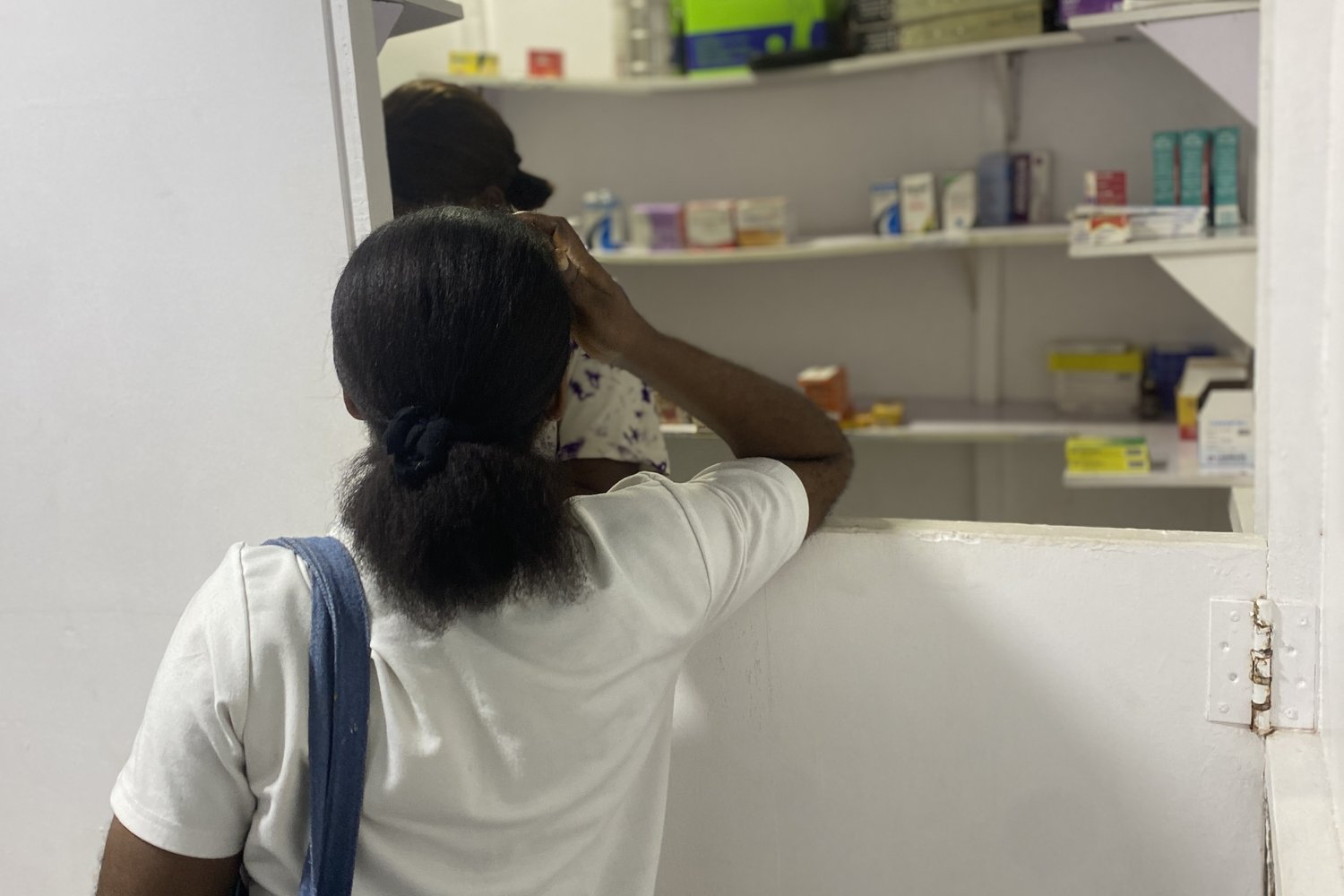 Woman with a blue tote bag standing at a pharmacy counter while a staff member retrieves medication from white shelves.