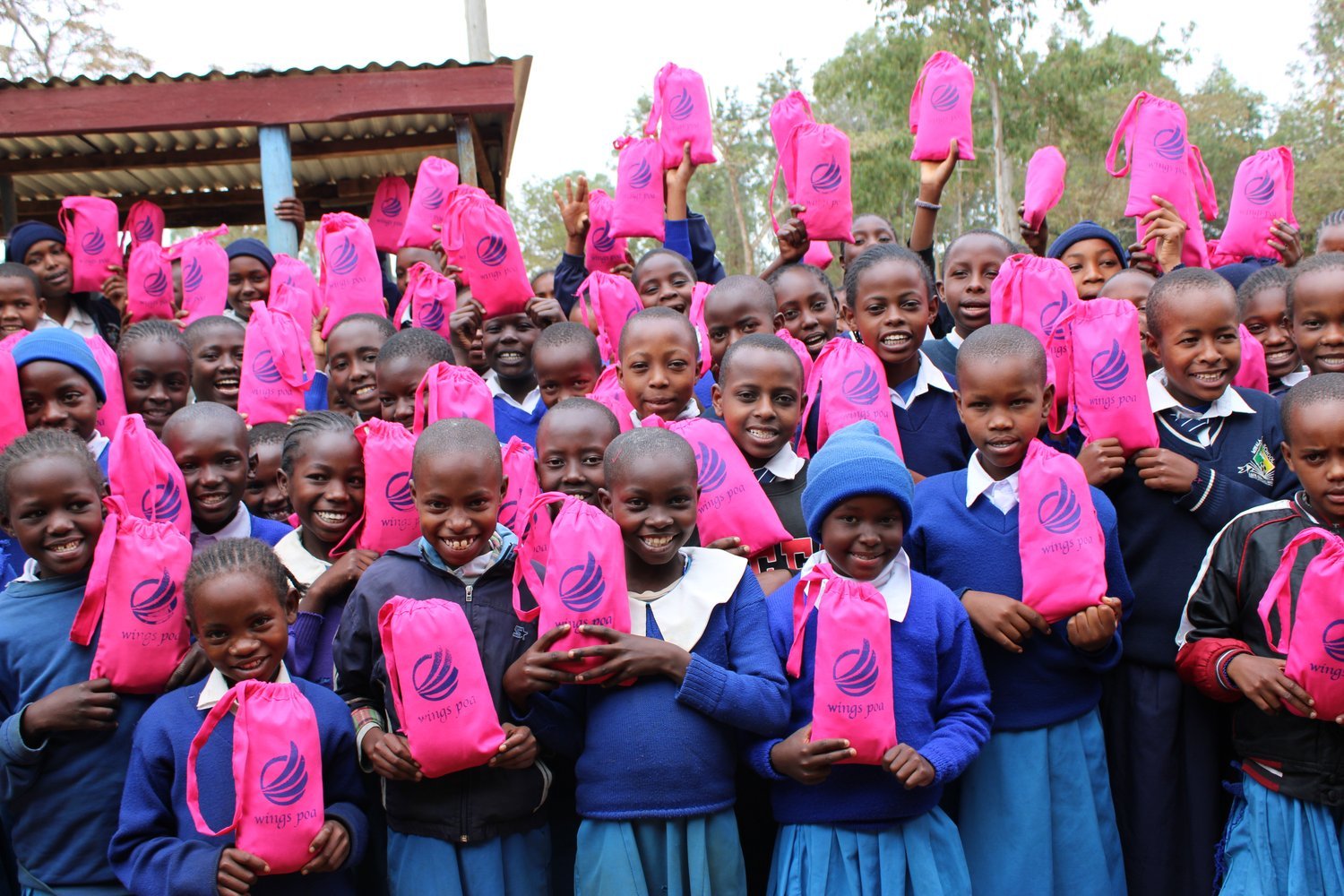 Children in blue school uniforms holding pink branded drawstring bags outdoors near a building with a corrugated metal roof.