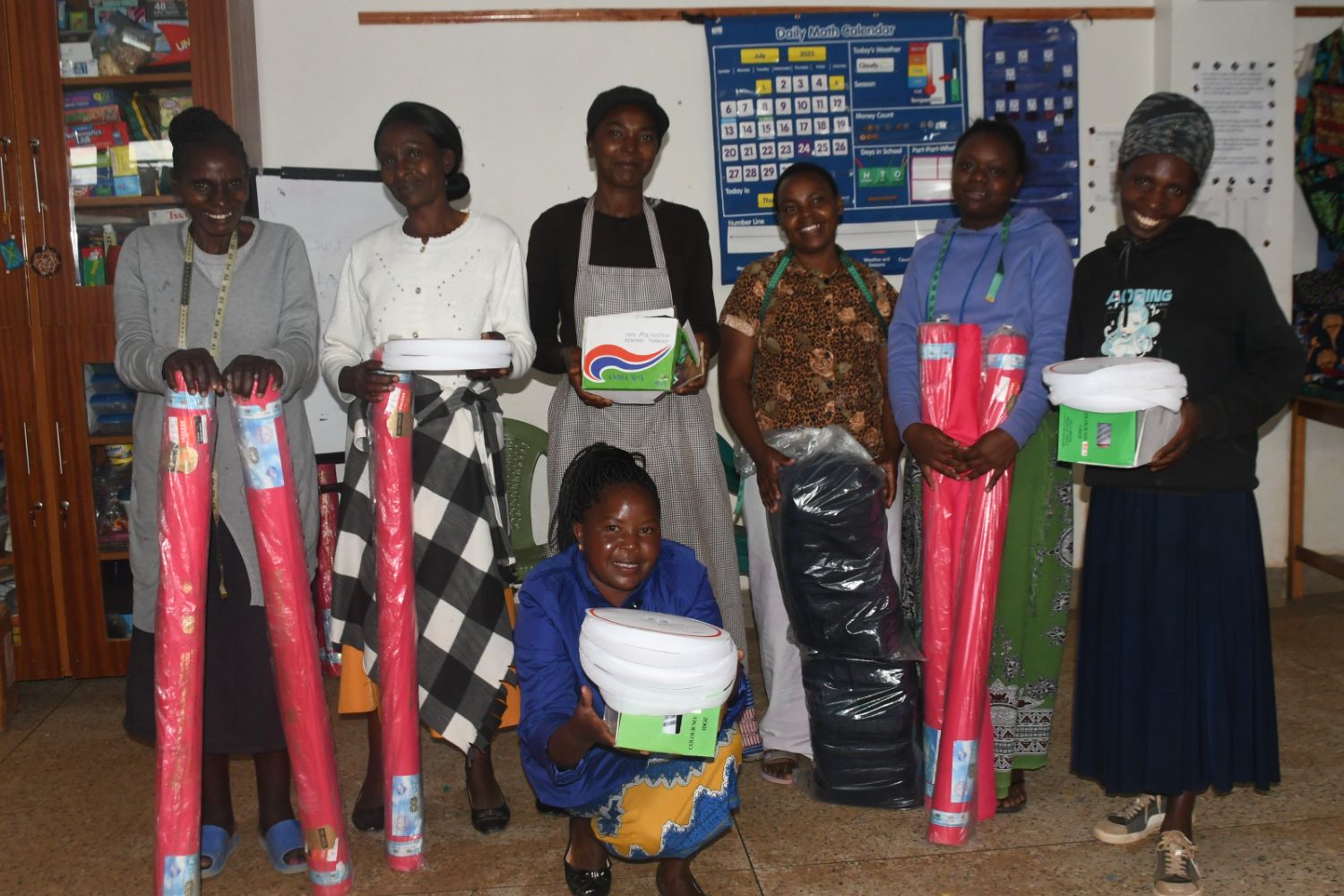 Seven women smiling, holding colorful rolls of fabric and boxes, standing indoors with a calendar on the wall behind them.