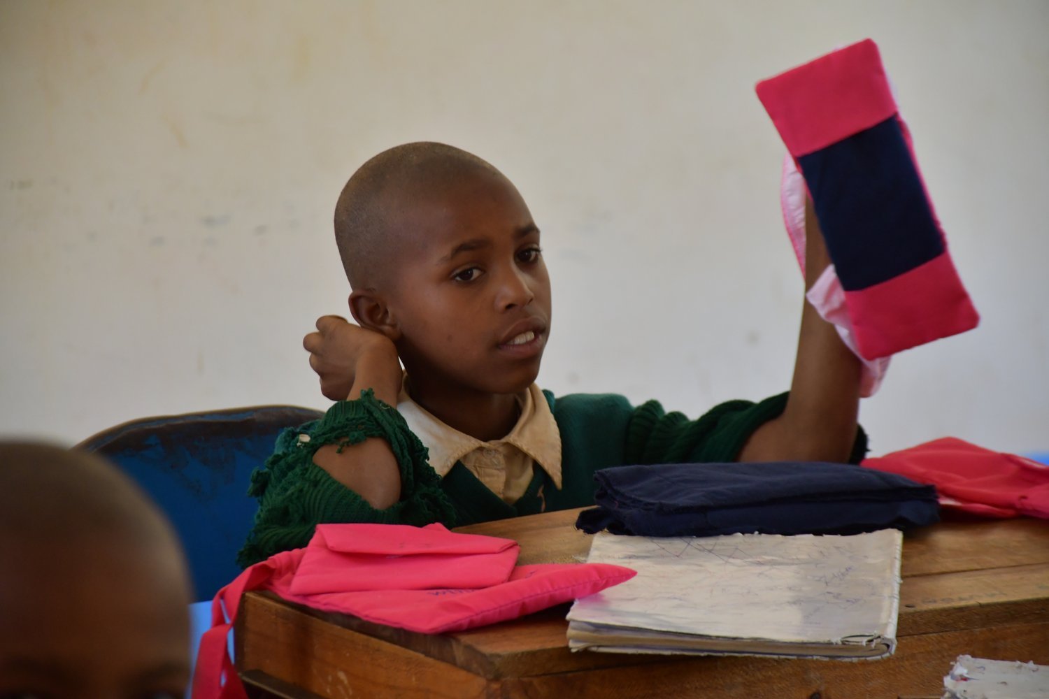 A young student in a green school uniform holds up a red and navy item at a wooden classroom desk.