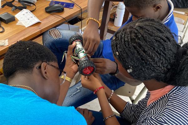 Three students assembling a small motorised wheel component with wiring in a classroom.
