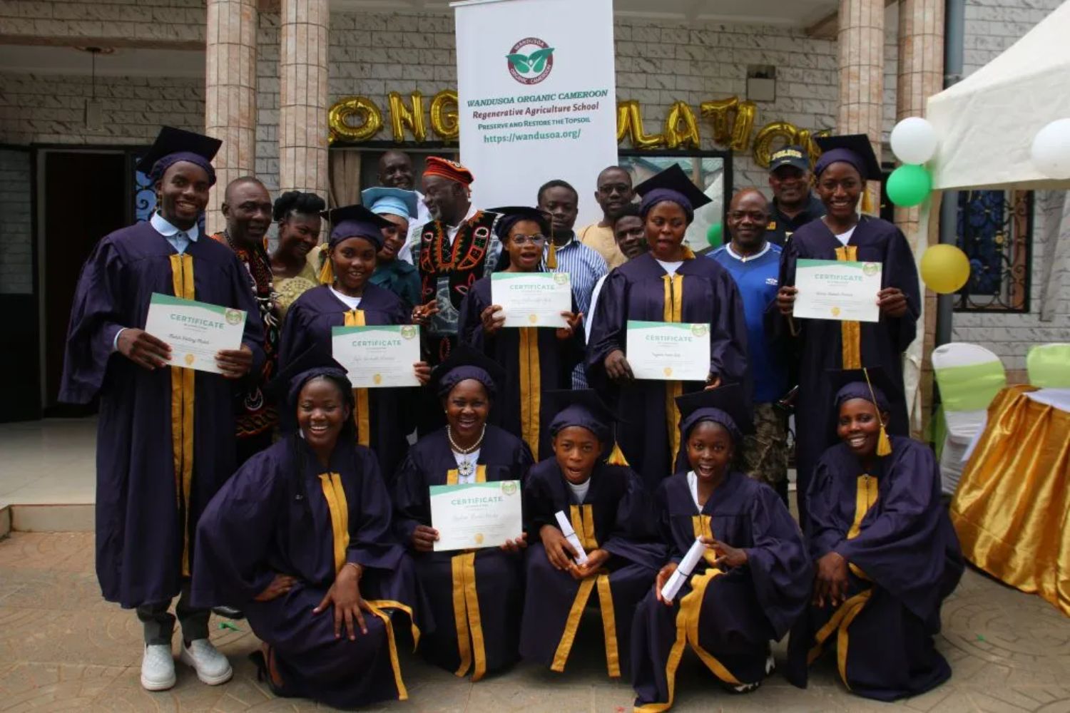 Graduates in caps and gowns proudly hold certificates, posing with faculty and family in front of a decorated venue.
