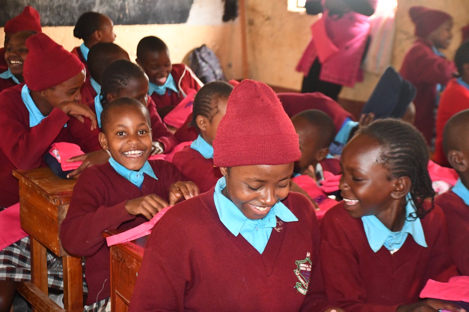 Children in maroon and blue school uniforms sitting at wooden desks in a classroom.
