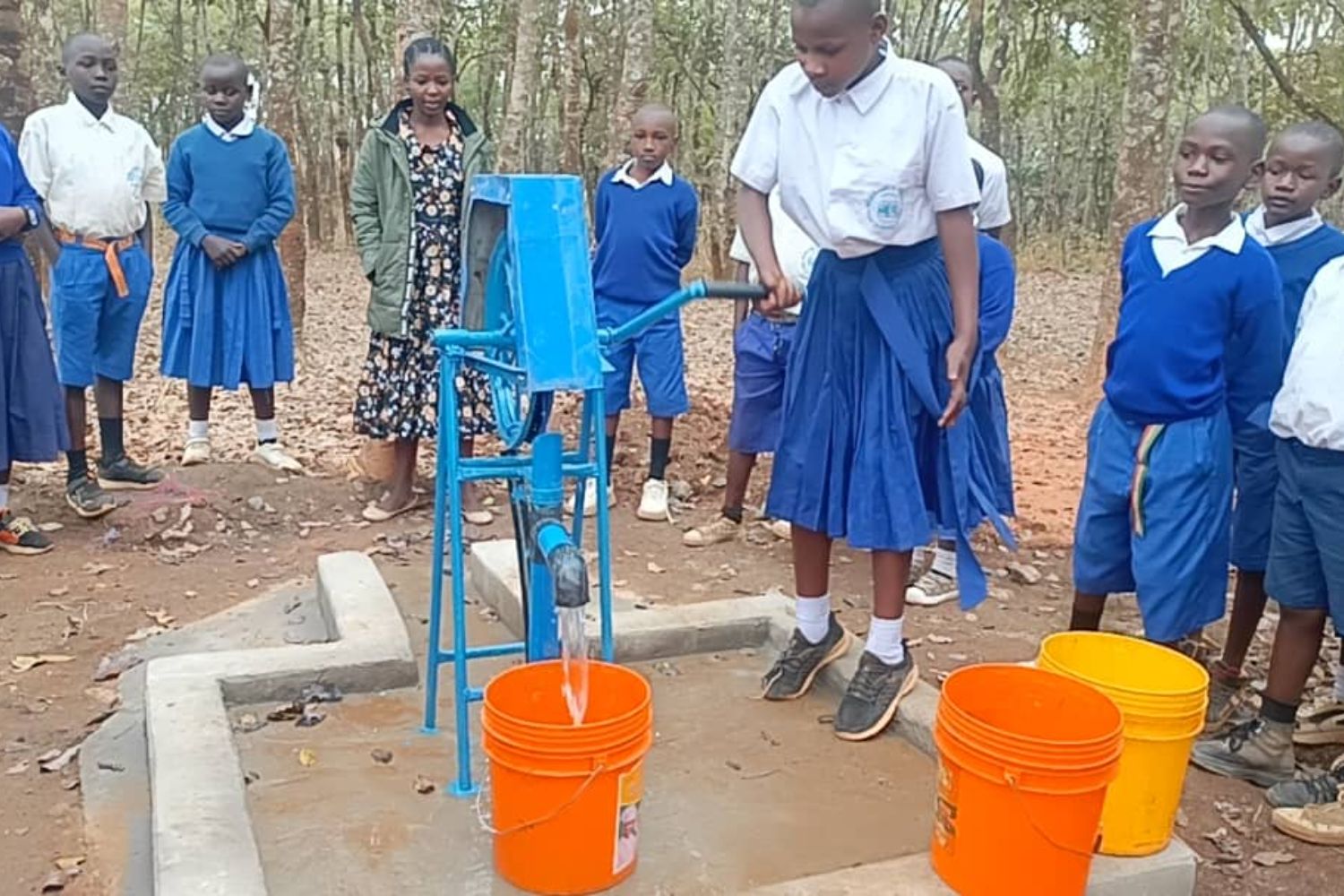 A group of students in uniforms gather around a blue water pump. One student operates it, filling orange buckets with water.