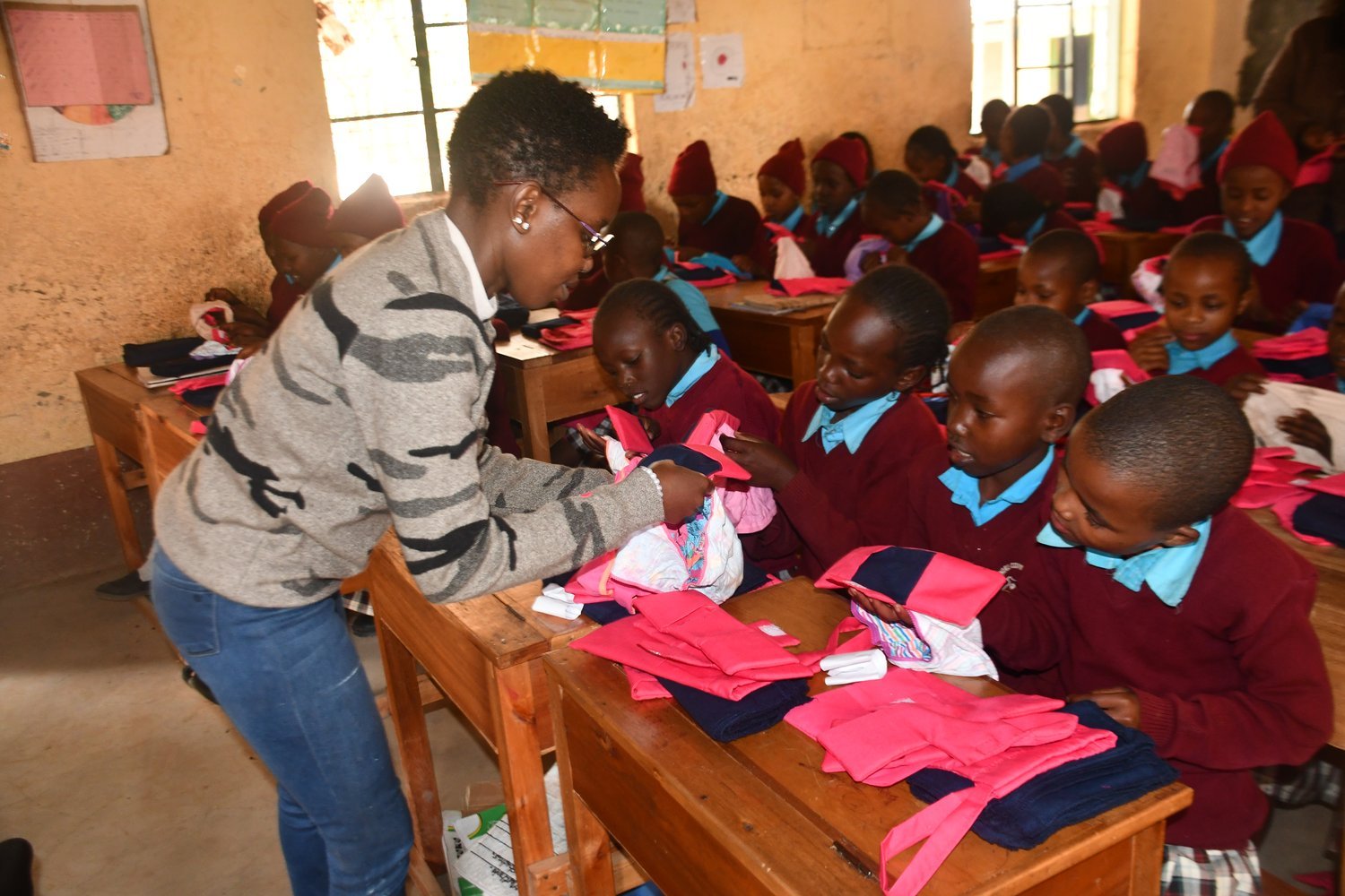 A woman in a grey sweater distributing pink and navy items to children in burgundy and blue uniforms at wooden school desks.