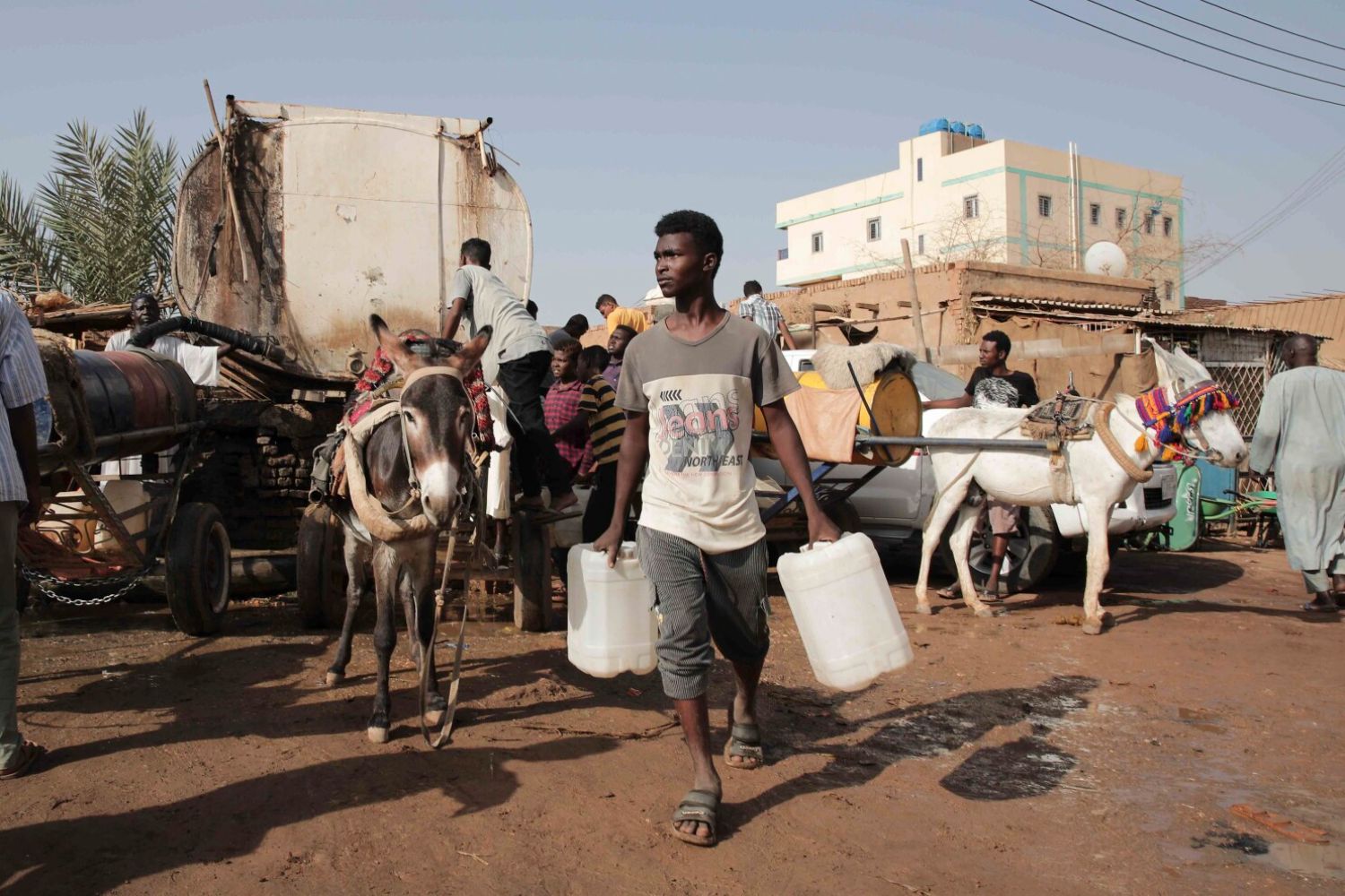 A man carries water containers in a busy street with donkeys and carts, while others work around him under a clear sky.