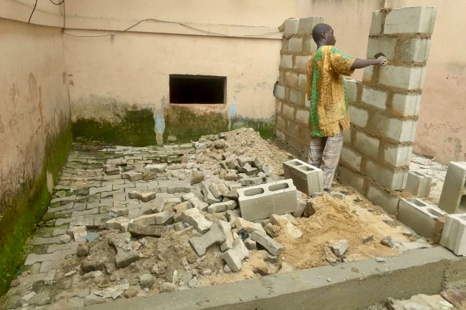 A person building a wall with concrete blocks in a small, unfinished courtyard area. Debris and sand are scattered on the ground.