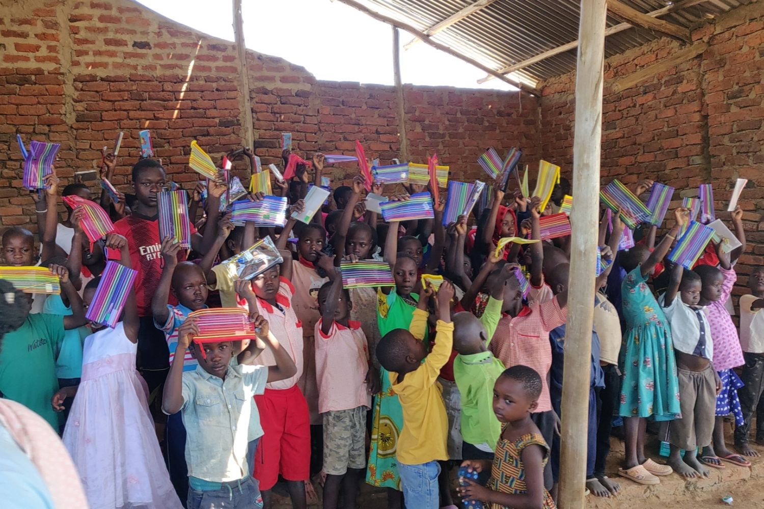 Children in a rustic classroom hold up colorful notebooks, smiling and standing closely together against a brick wall under a metal roof.