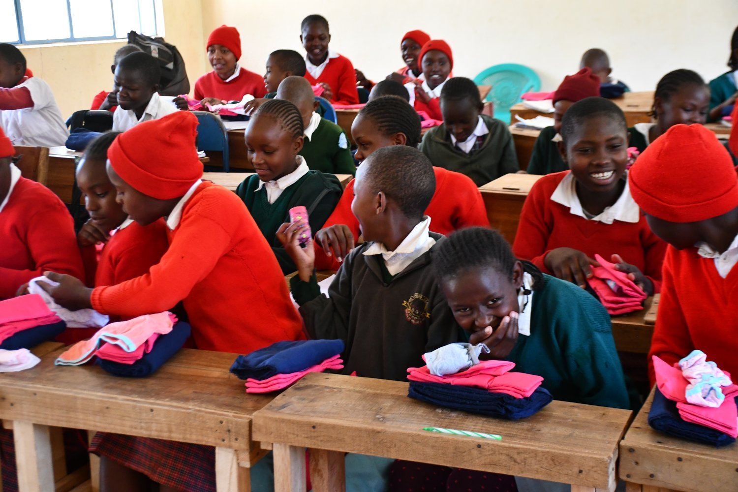 Children in red and green uniforms examining folded clothing items at wooden desks in a classroom.