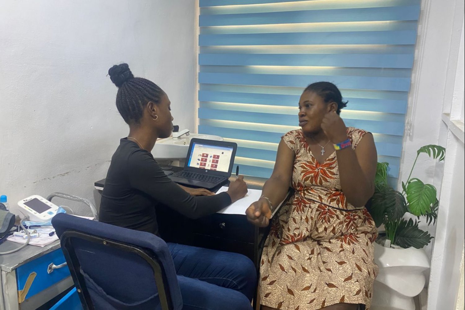 Woman in a patterned dress speaking with a healthcare worker at a desk with a laptop in a medical consultation room.