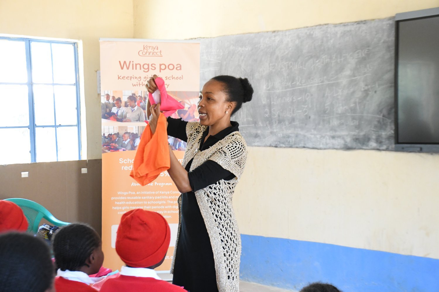 A woman holds up colourful reusable sanitary pads while presenting the Kenya Connect Wings Poa programme to students in a classroom.