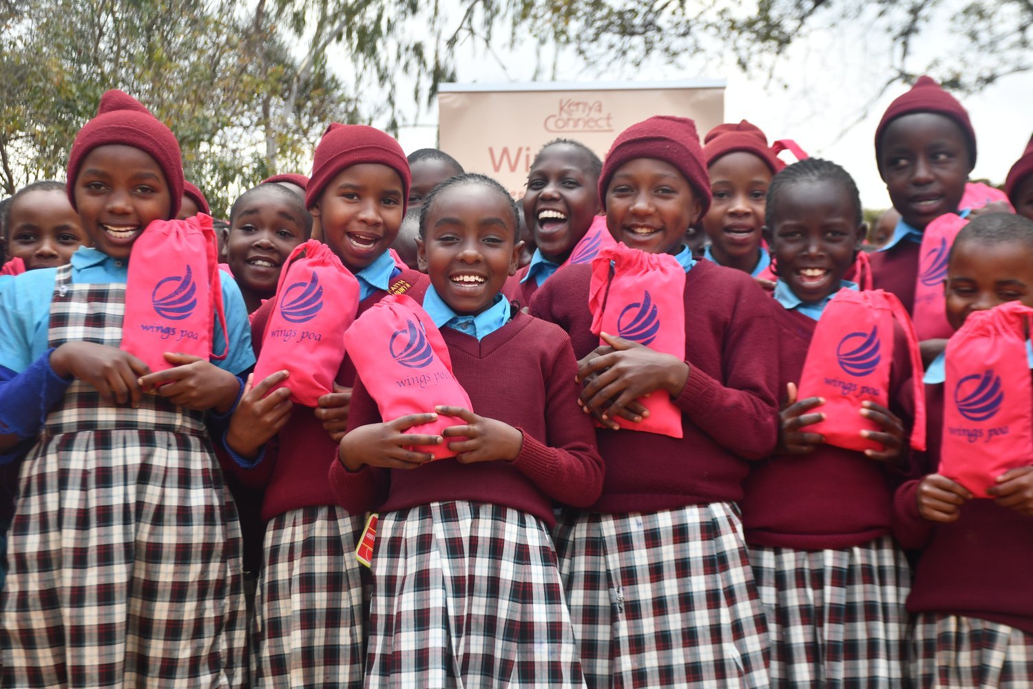 Girls in maroon school uniforms holding pink "Wings Poa" branded bags outdoors, with a "Kenya Connect" banner visible behind them.