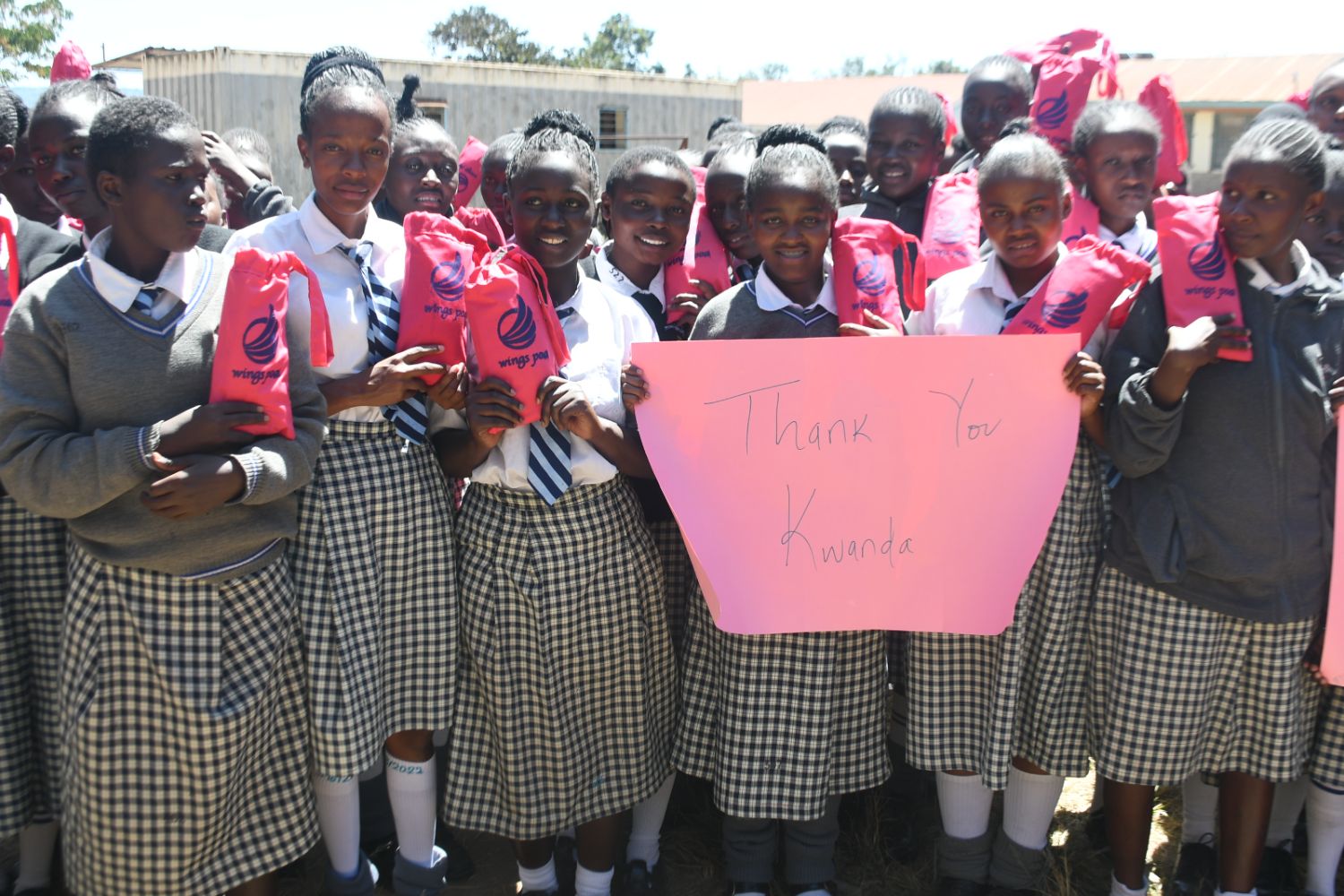 A group of schoolgirls in uniforms hold reusable menstrual kits and a pink sign reading "Thank You Kwanda" outdoors.