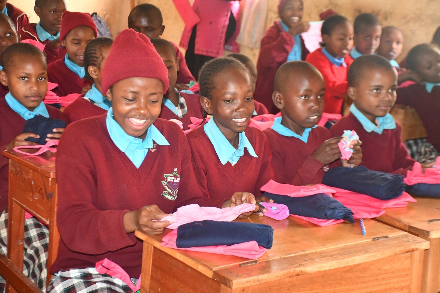 Children in maroon and blue uniforms sitting at wooden desks holding pink and navy clothing items in a classroom.