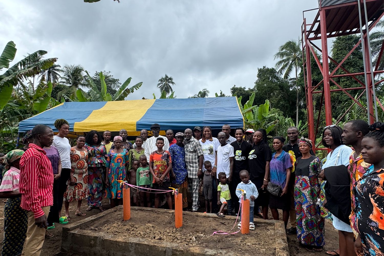 A diverse group of people stands together at a construction site with a colorful tent and tropical trees in the background.