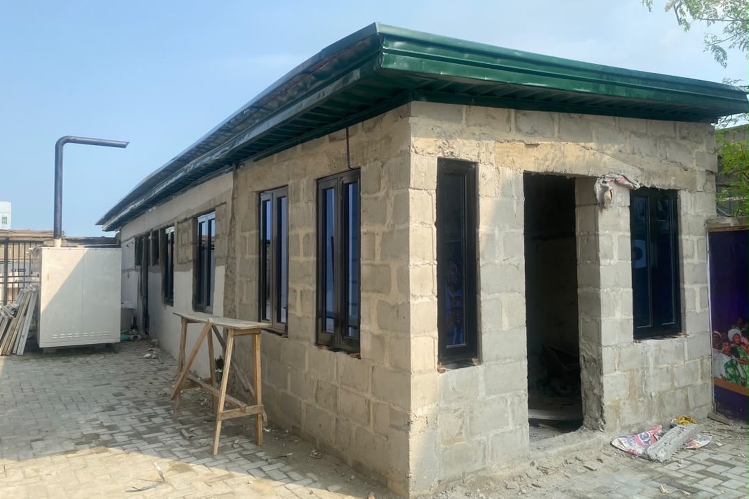 A single-storey concrete block building under construction with a green metal roof and dark-framed windows on a paved courtyard.
