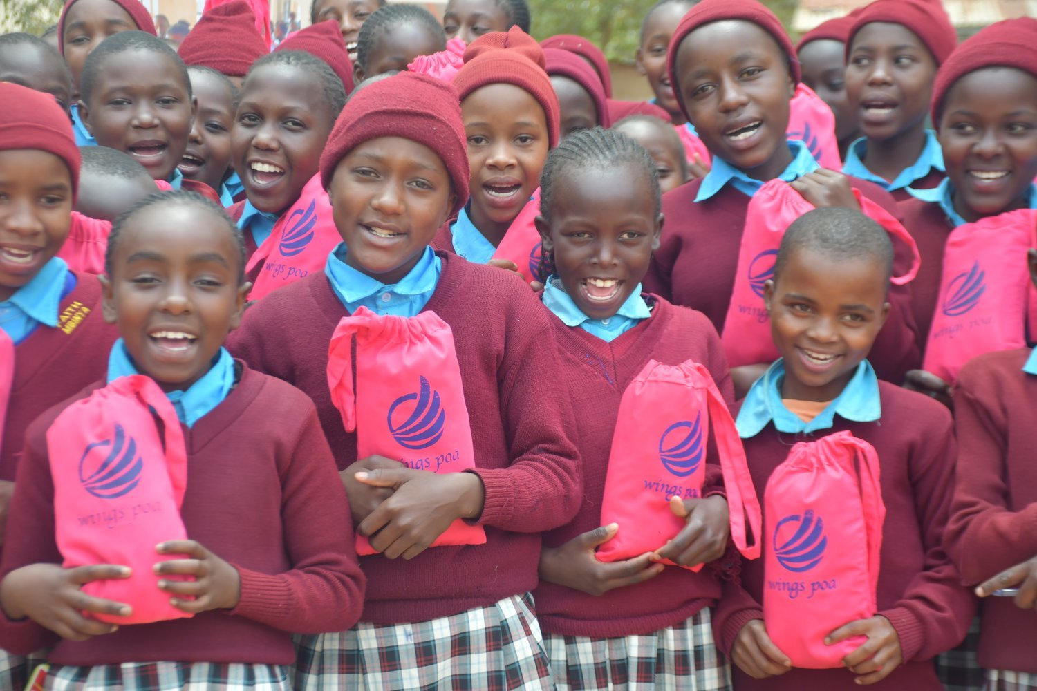 Girls in maroon school uniforms holding pink "Wings Poa" branded bags outdoors in a group setting.