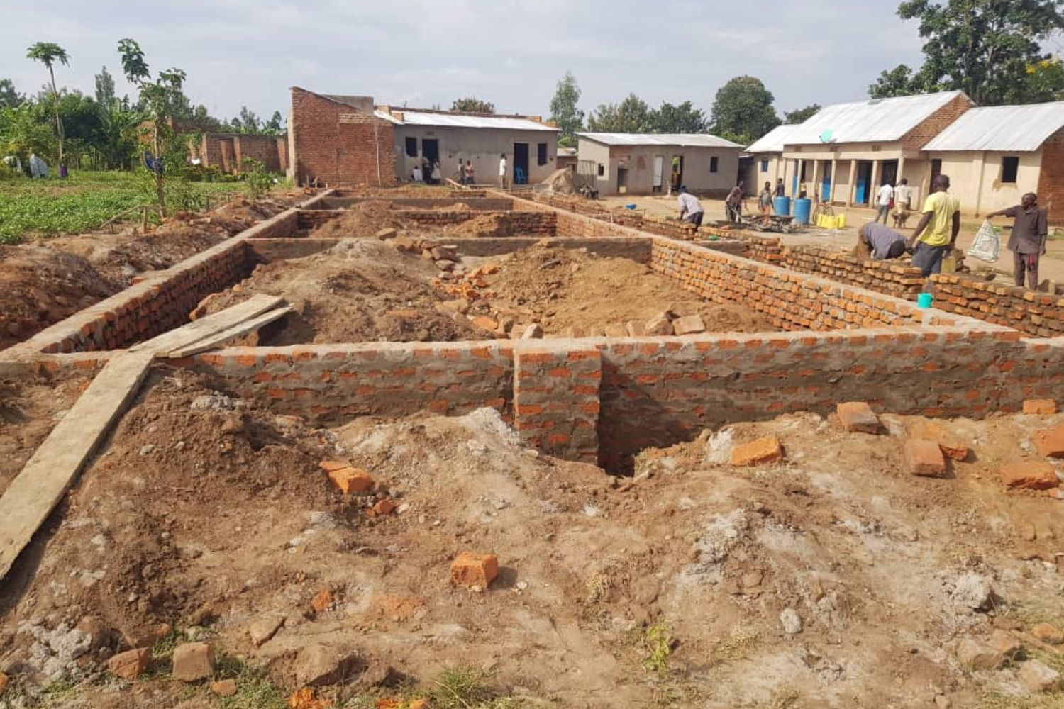 Construction site with brick foundation walls, surrounded by dirt and debris. Several people are working near small buildings in the background.
