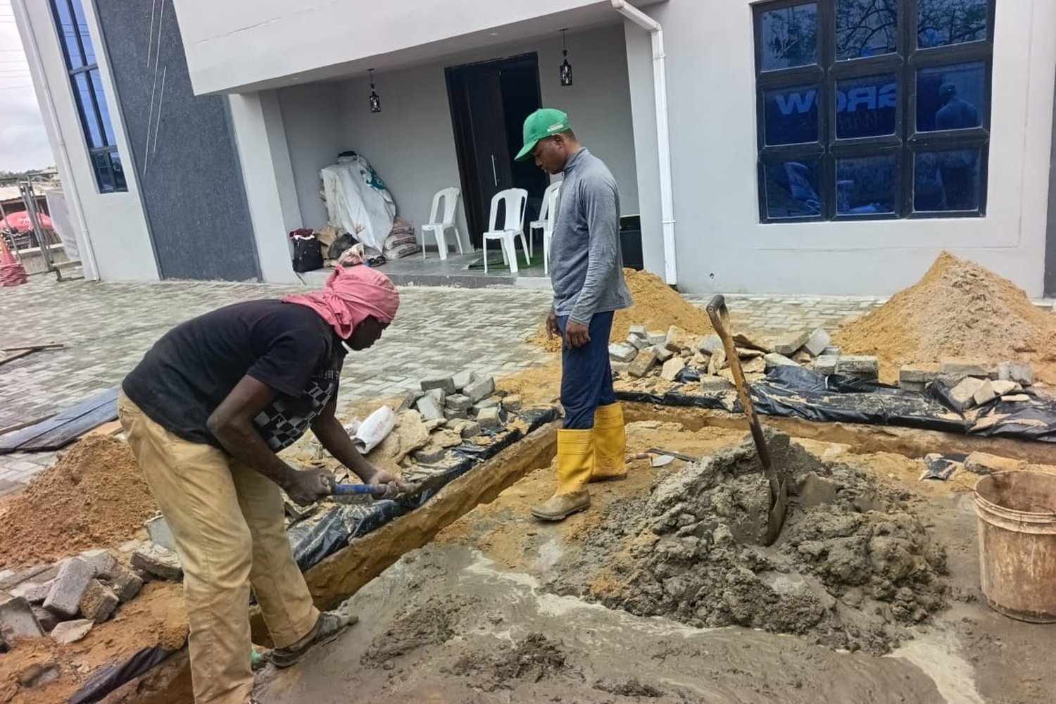 Two construction workers laying paving stones and concrete in a trench outside a modern grey building with a covered entrance.