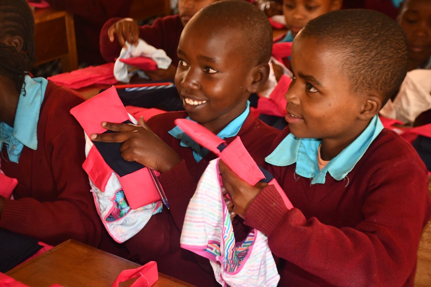 Children in maroon and blue school uniforms handling pink bags and clothing items whilst seated at wooden desks in a classroom.
