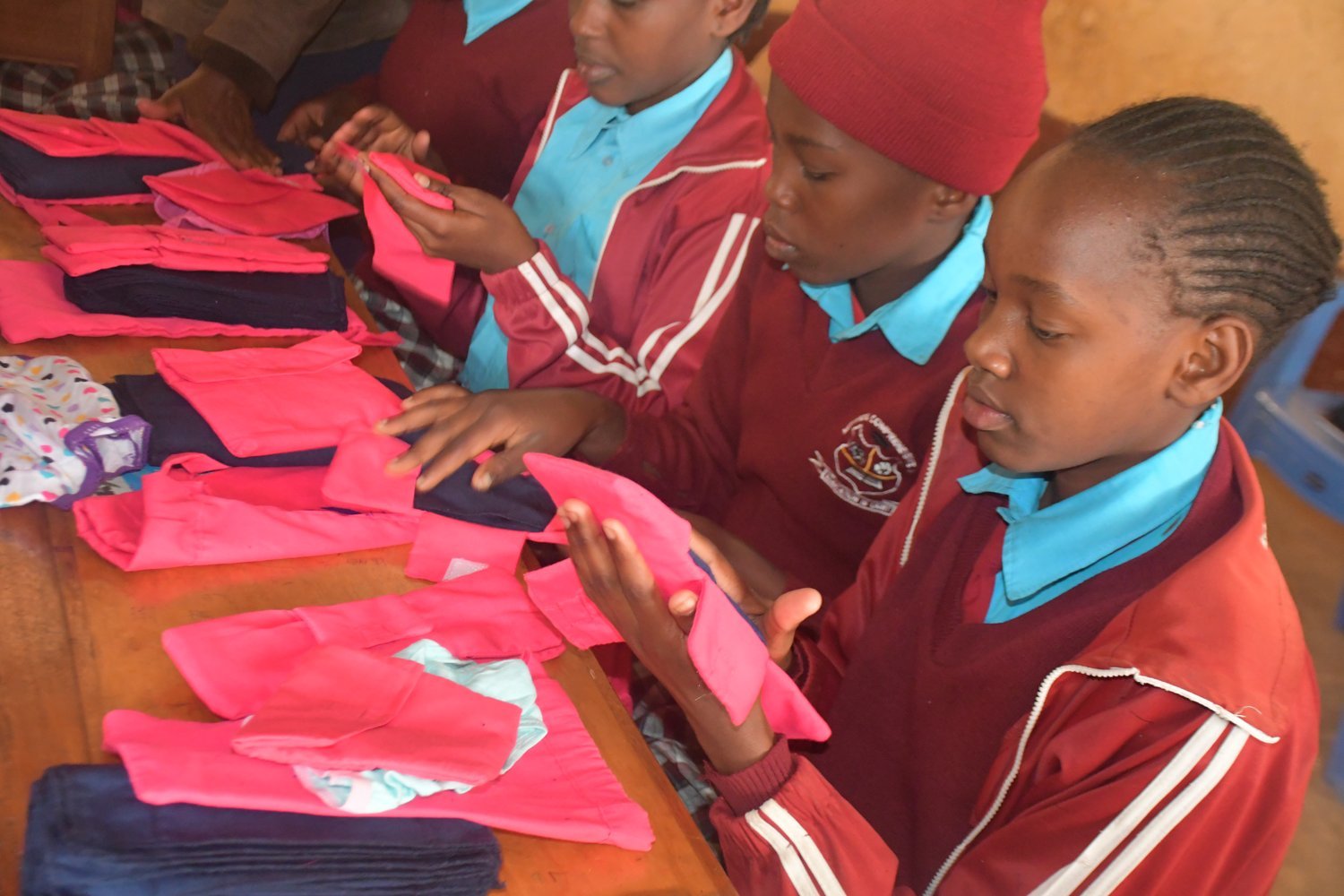 Children in maroon and blue school uniforms examining pink and navy fabric items on a wooden table indoors.