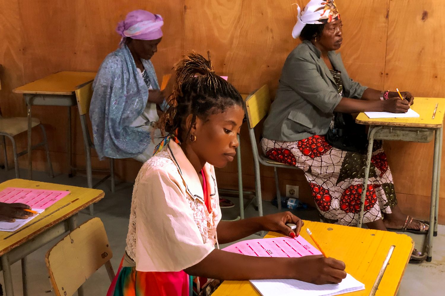 Women seated at desks in a classroom, writing in notebooks. They wear colorful clothing and headscarves, with wooden walls in the background.
