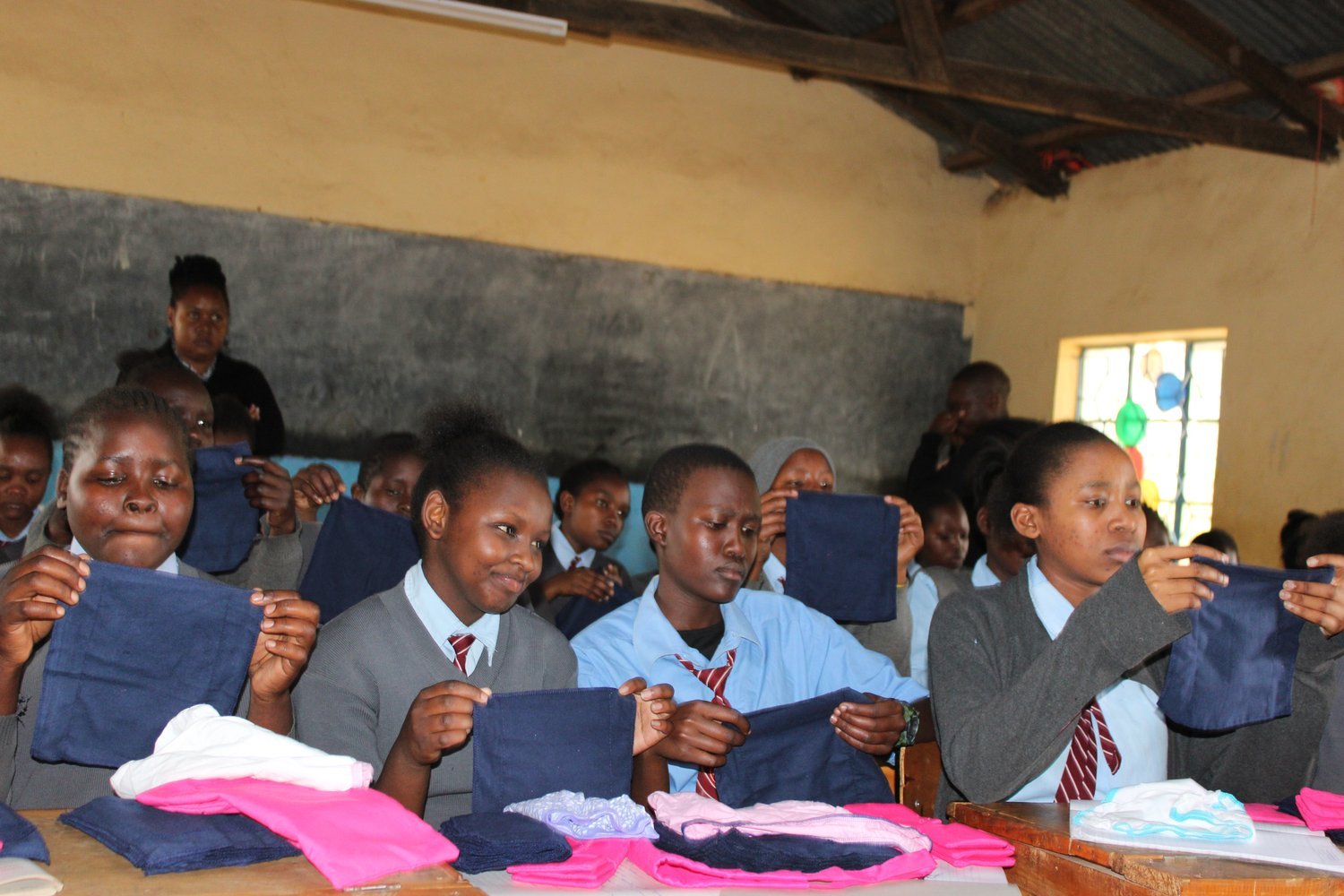 Students in grey blazers and blue uniforms holding dark navy fabric items in a classroom with a chalkboard.
