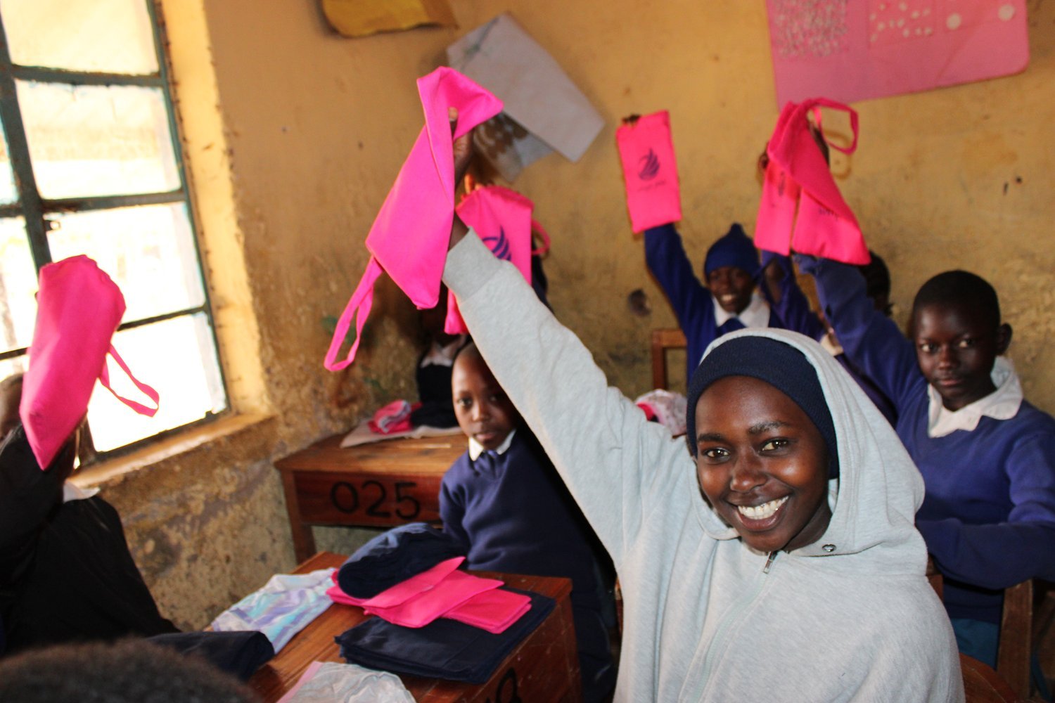 Students in blue uniforms holding up bright pink bags in a classroom with yellow walls and wooden desks.