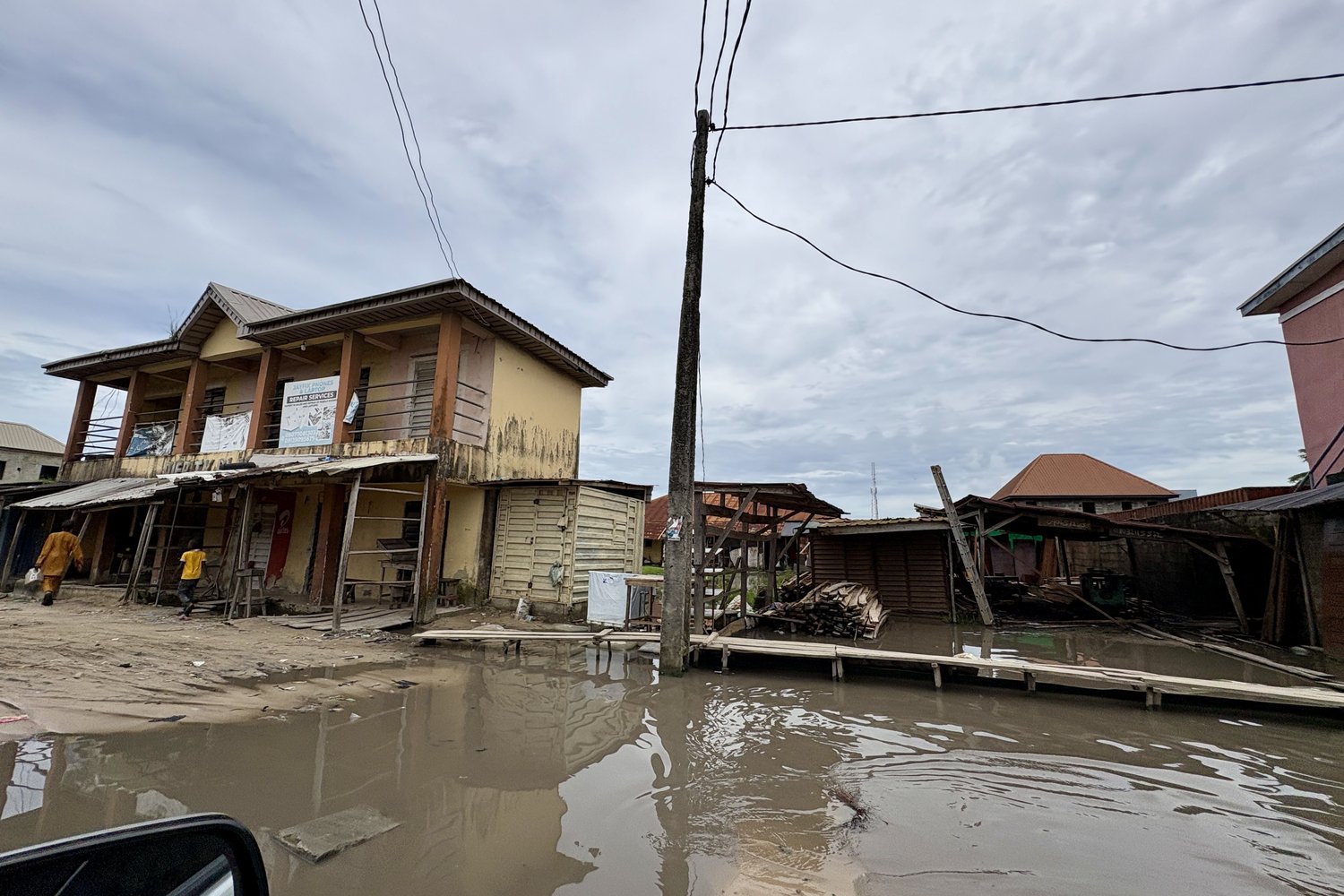 A waterlogged sand road through Onosa, lined with single-storey shopfronts and a two-storey building