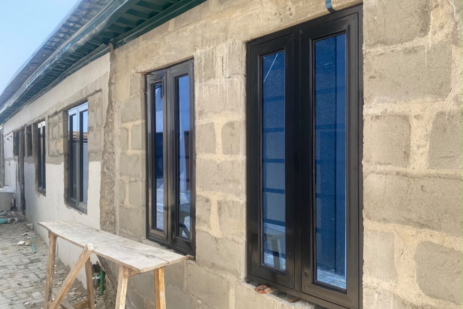 A building under construction with dark-framed glass windows installed in an unpainted concrete block wall beside a wooden workbench.