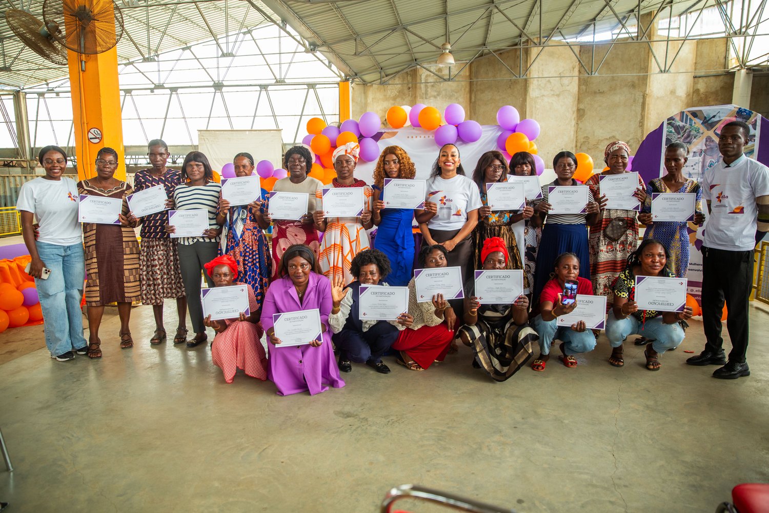 About twenty women standing in a hall holding participation certificates, under orange and purple balloons.