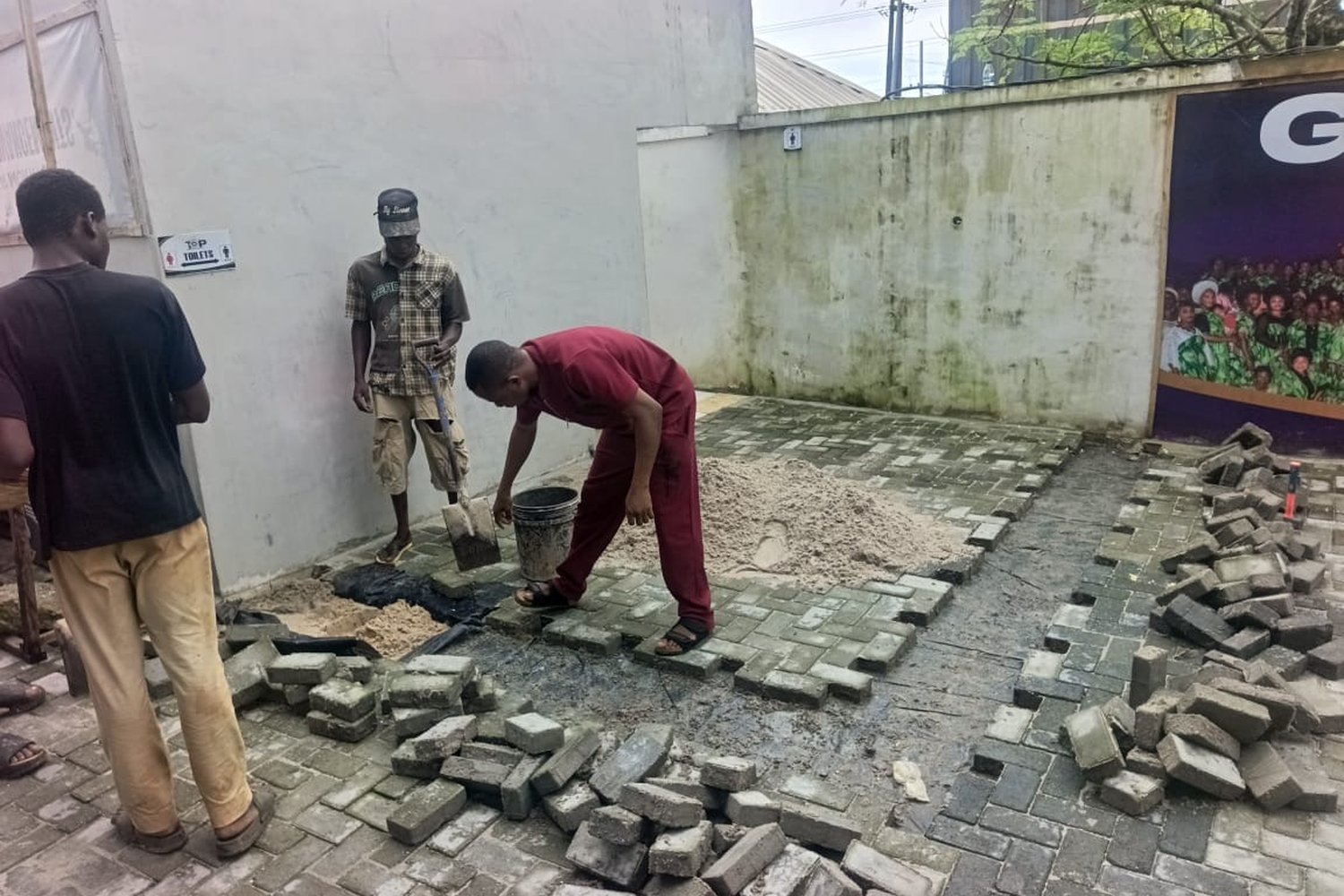 Three men laying grey concrete paving blocks on a courtyard floor beside a white rendered wall, with sand and a bucket nearby.