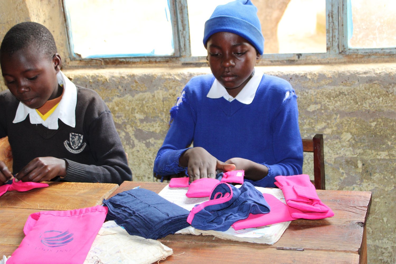 Two schoolchildren in uniforms sewing pink and blue fabric at wooden desks in a classroom with peeling walls.