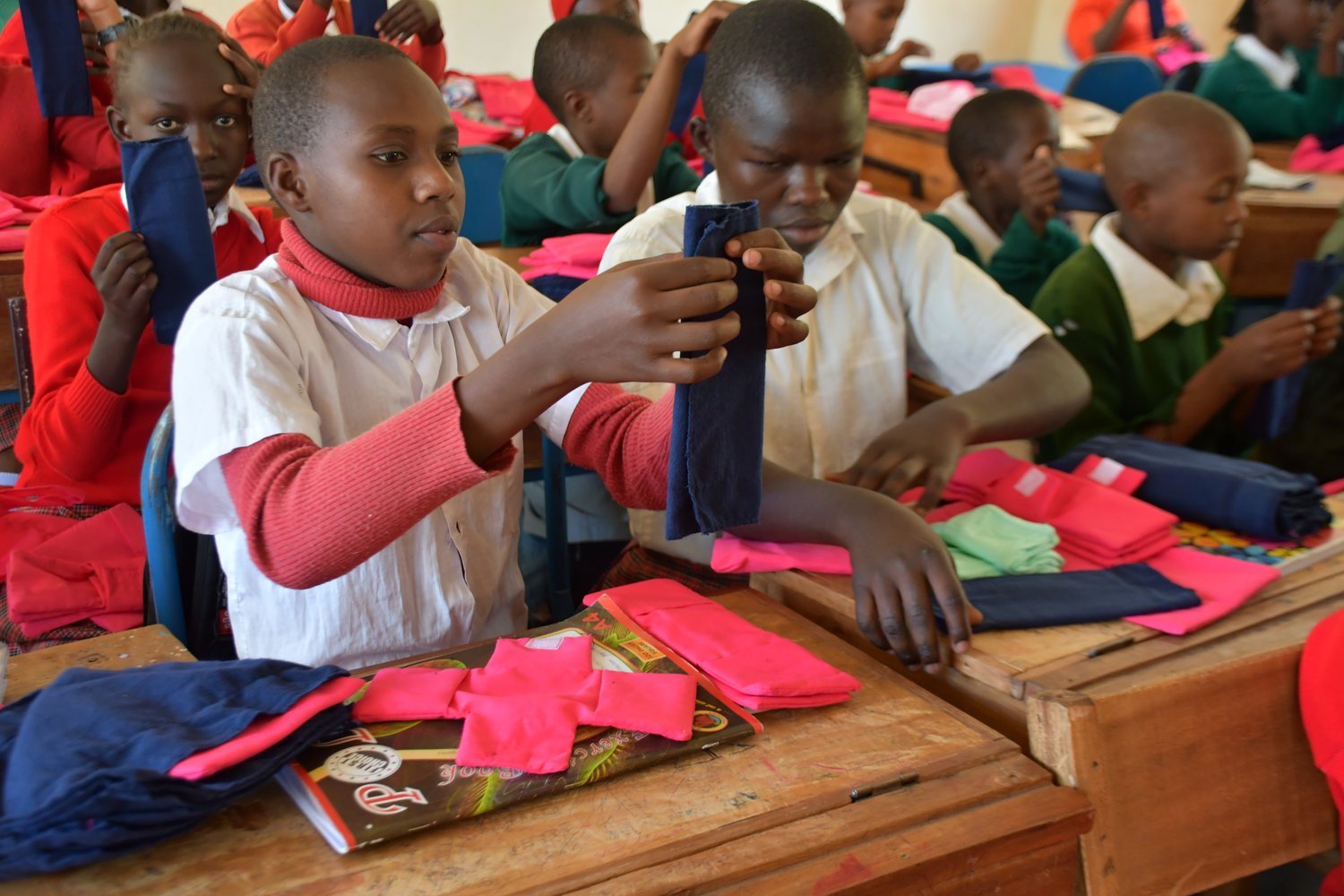 Children in school uniforms examining pink and navy blue fabric items at wooden desks in a classroom.
