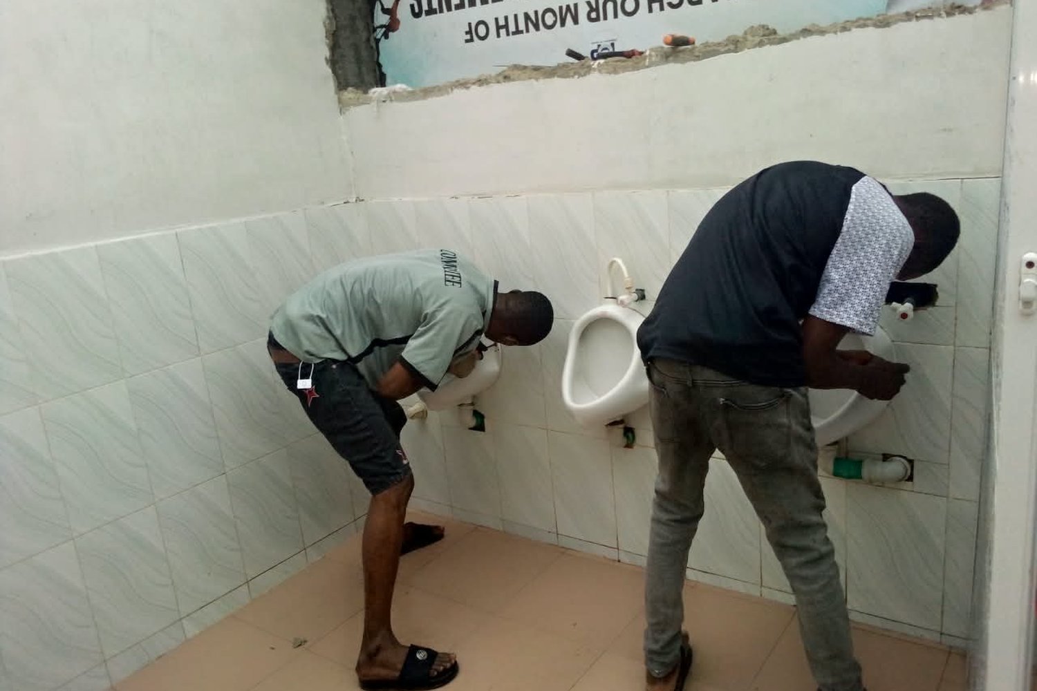 Two men installing wall-mounted urinals in a tiled public bathroom with white walls and a partially demolished upper section.