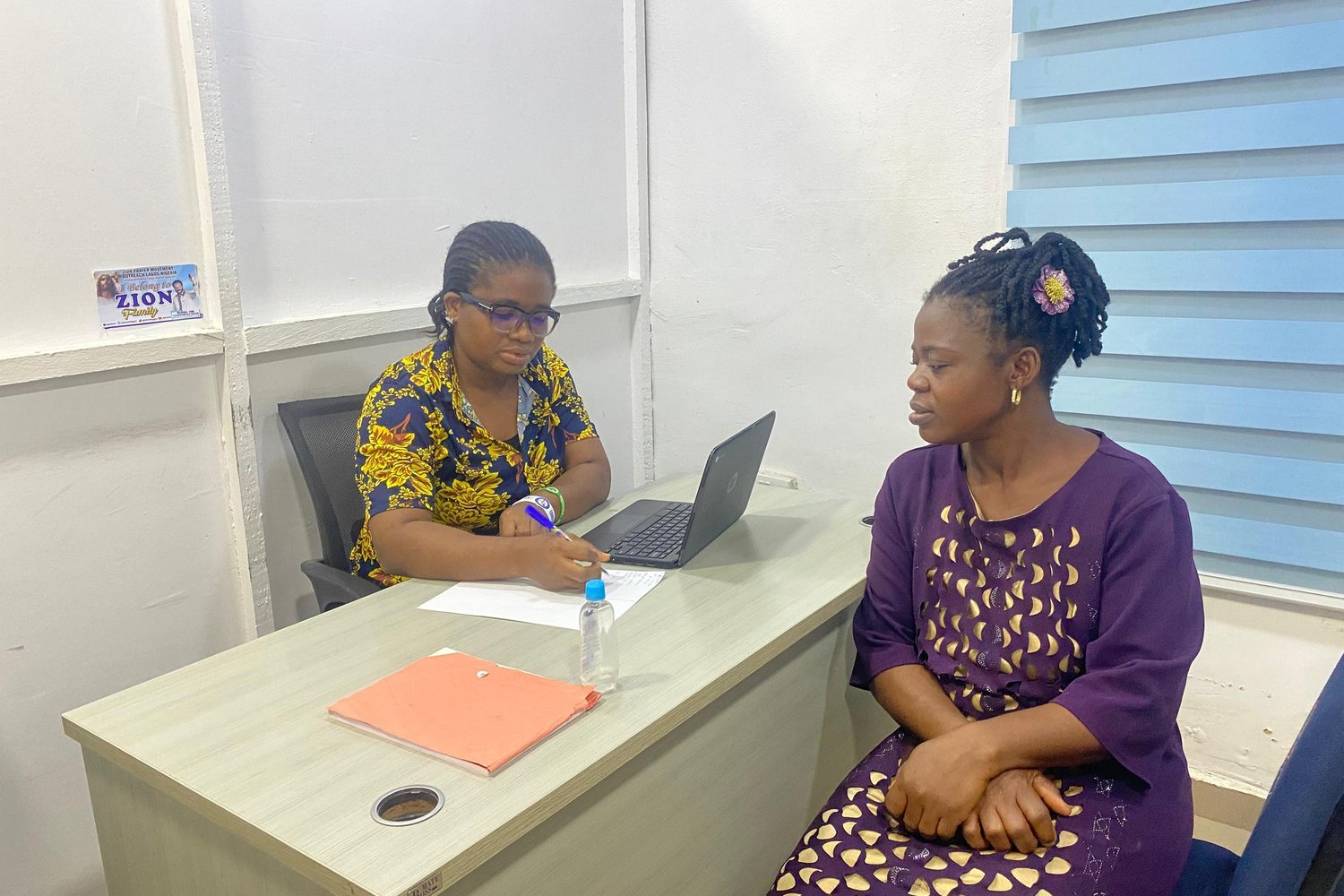 Two women in colourful clothing sitting at an office desk with a laptop, one taking notes while the other faces her.