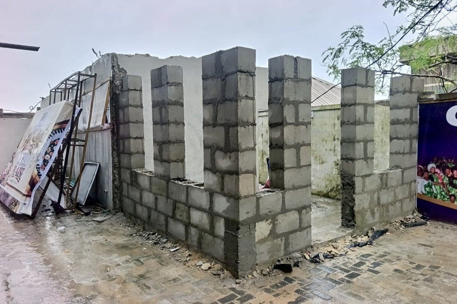 Concrete block pillars under construction beside a partially demolished grey wall on a wet paved courtyard.