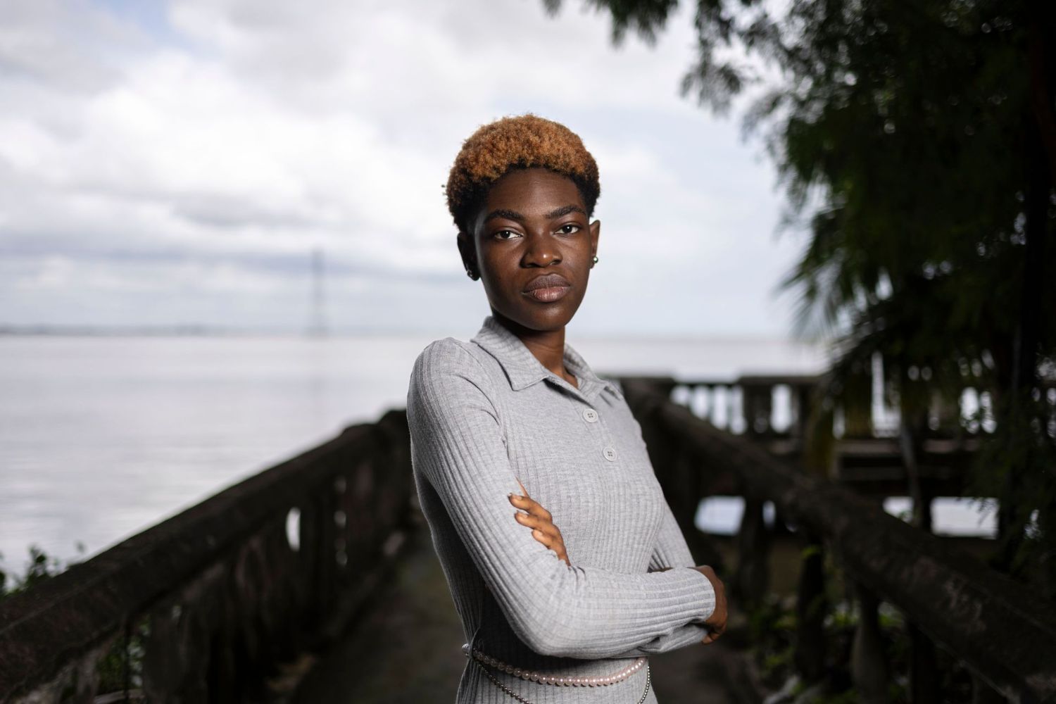 Person in a gray sweater stands confidently with crossed arms on a wooden pathway near a body of water, surrounded by trees.