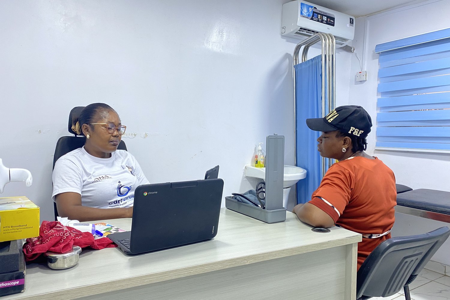 A healthcare worker in a white T-shirt consulting with a patient across a desk with a Lenovo Chromebook in a medical centre.