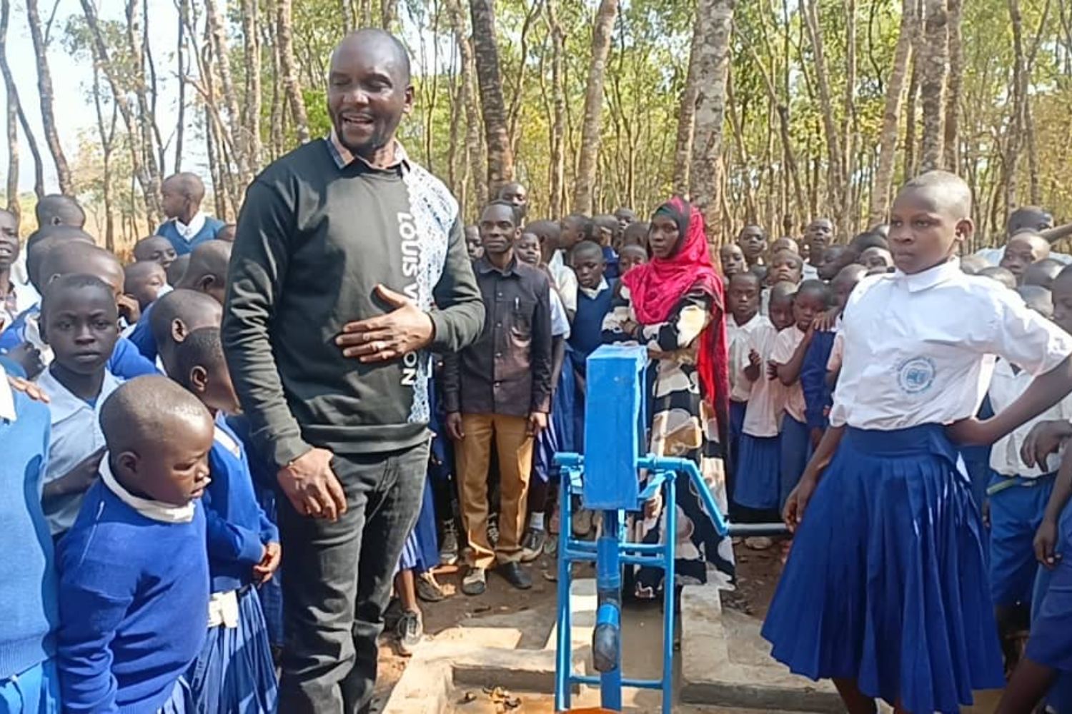 A man stands beside a water pump surrounded by schoolchildren in blue uniforms, with trees in the background.
