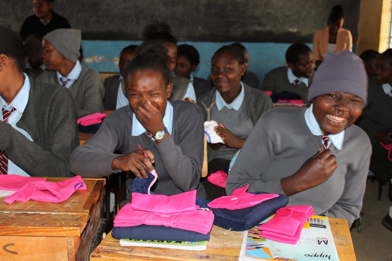 Students in grey uniforms examining folded pink and navy garments on wooden desks in a classroom with a chalkboard.