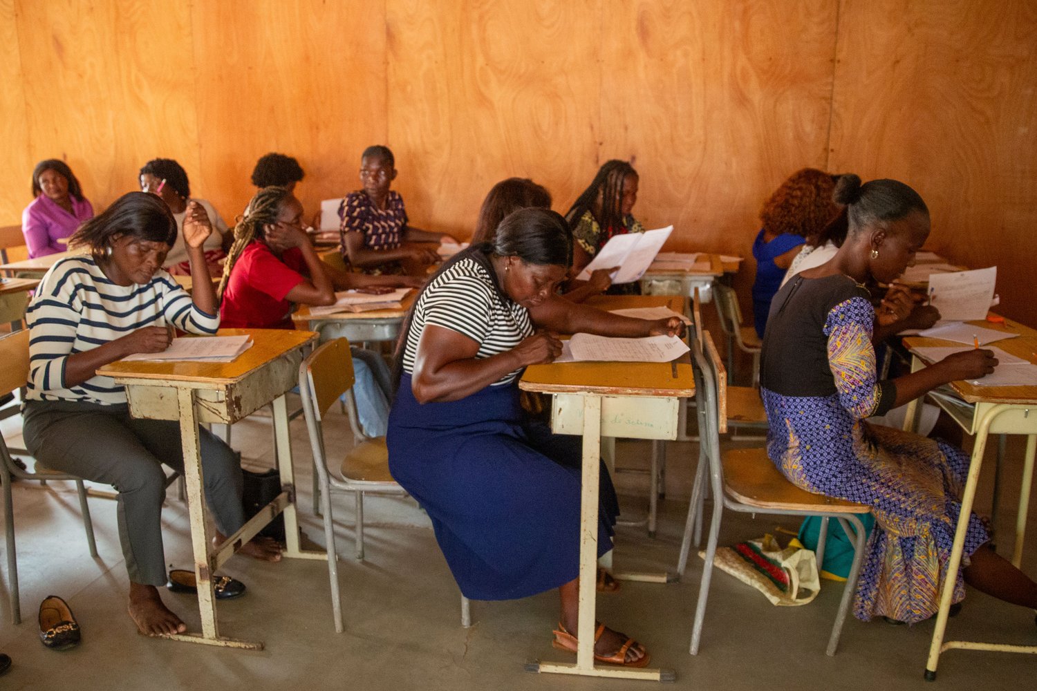Six women at wooden school desks reading and annotating printed worksheets in a plywood-walled classroom.