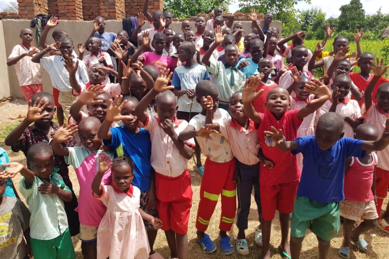 A large group of smiling children outdoors, waving enthusiastically at the camera. They are dressed in colorful clothing.