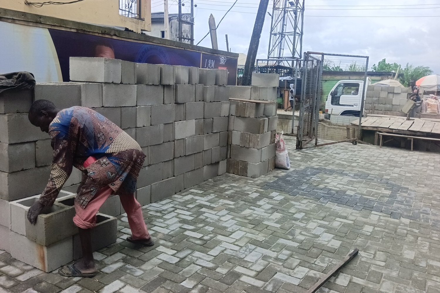 A worker in a patterned shirt laying grey concrete blocks to build a low wall on a paved courtyard.