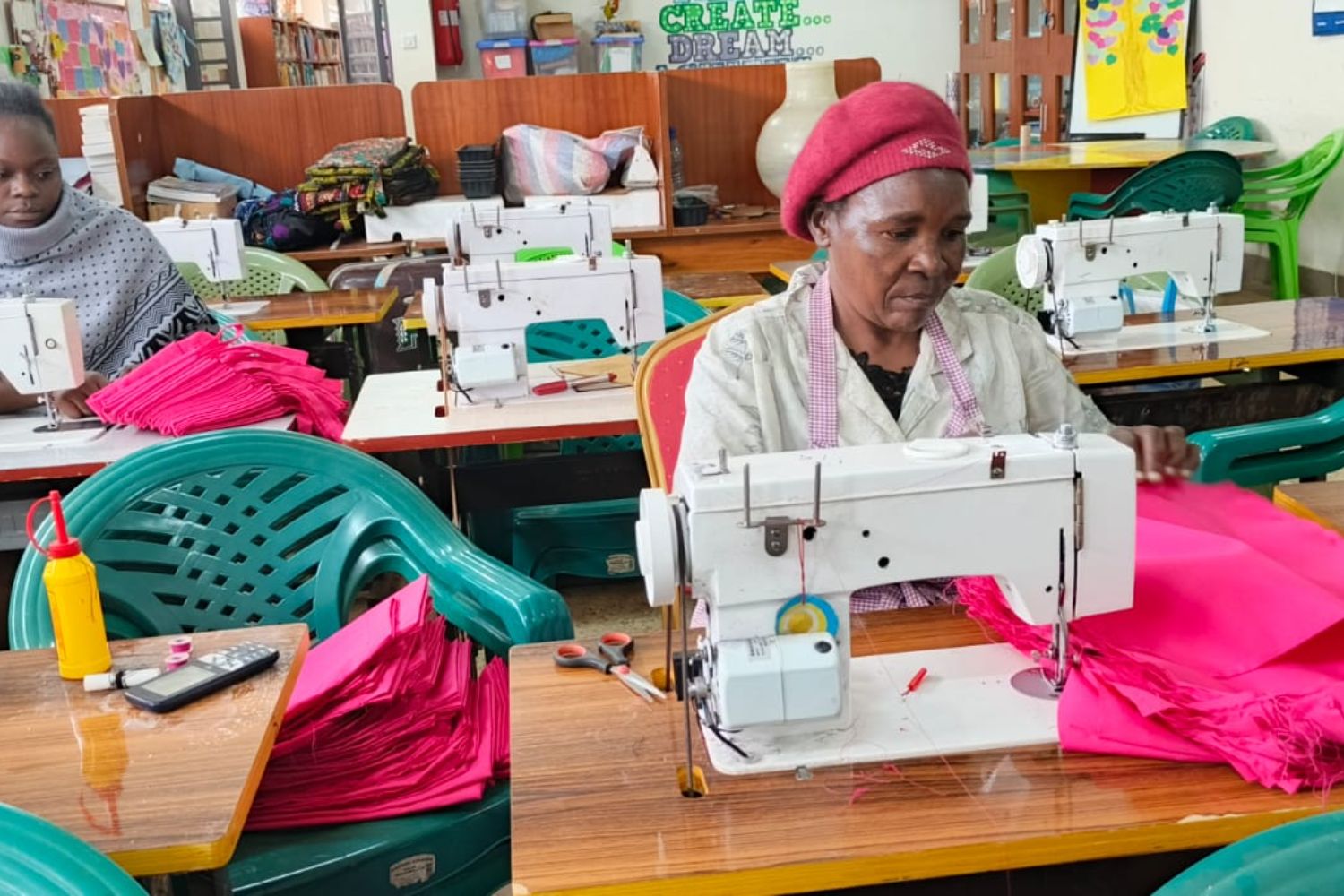Women sewing pink fabric on sewing machines in a workshop, surrounded by colorful chairs and tables with sewing materials.