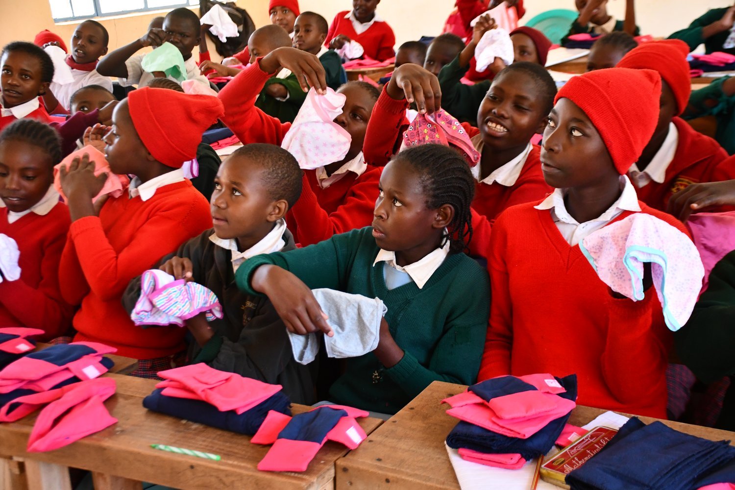 Children in red uniforms holding underwear and examining folded clothing items distributed on desks in a classroom.